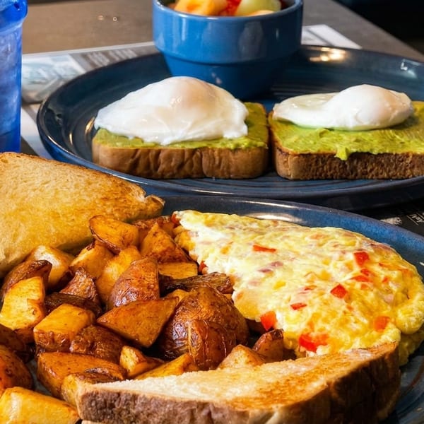 Two breakfast plates: one with omelet, potatoes, toast; another with avocado toast, poached eggs, fruit.