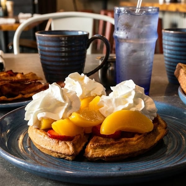 Breakfast spread with waffles topped with peaches and whipped cream, bacon, and toast.